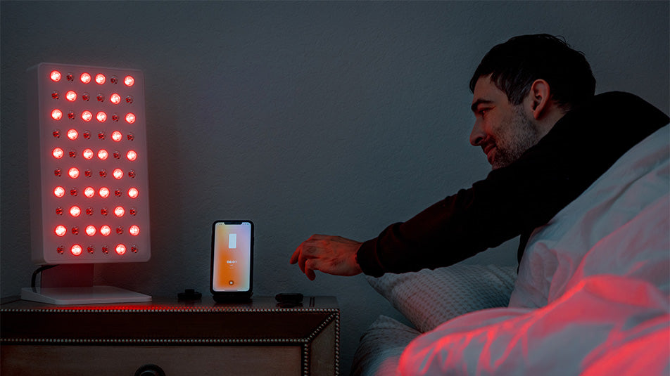 Man waking up in bed using the Joovv mobile app with Joovv Mini red light therapy panel as an alarm clock