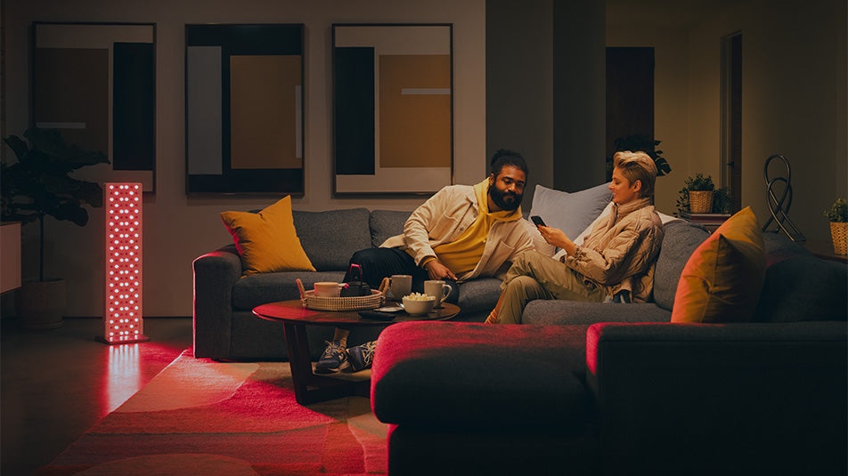 Two people relaxing on a couch using a Joovv light panel for ambient red light in a living room setting