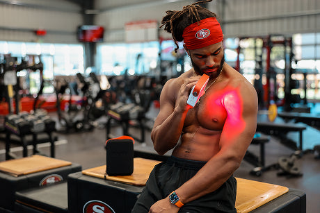 SF 49ers player using a JOOVV red light therapy Go 2.0 device on his arm in a gym setting